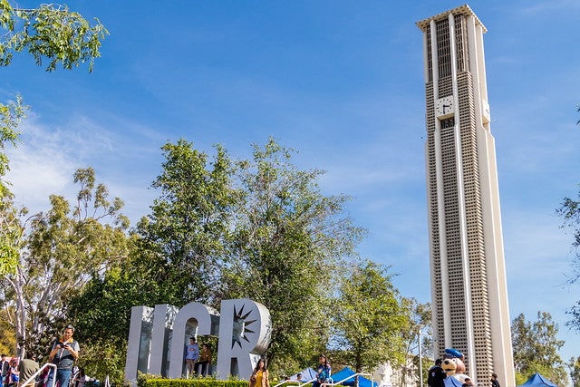 With the Bell Tower in the distance, a large metallic sculpture spells out UCR on the UC Riverside campus.