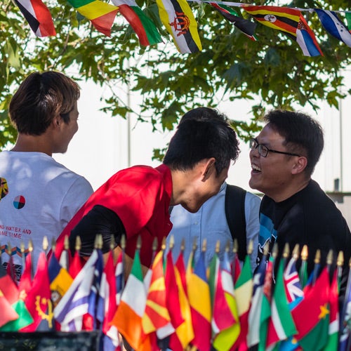 Three students share a laugh in front of a display of flags from many nations.